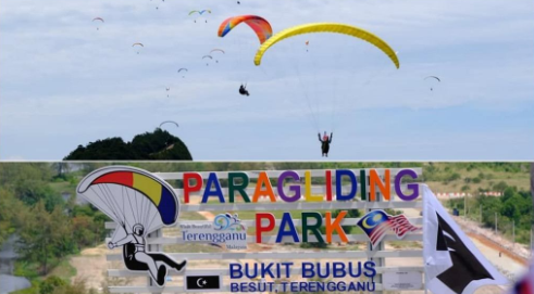 A large number of paragliders floating above an open field with a signboard saying "Paragliding Park Bukit Bubus"
